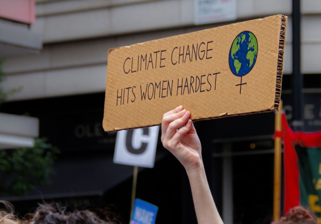 Hand håller handgjord skylt av kartong med texten "Climate change hits women hardest". Tillsammans med en ritad jordglob i ett kvinnotecken.